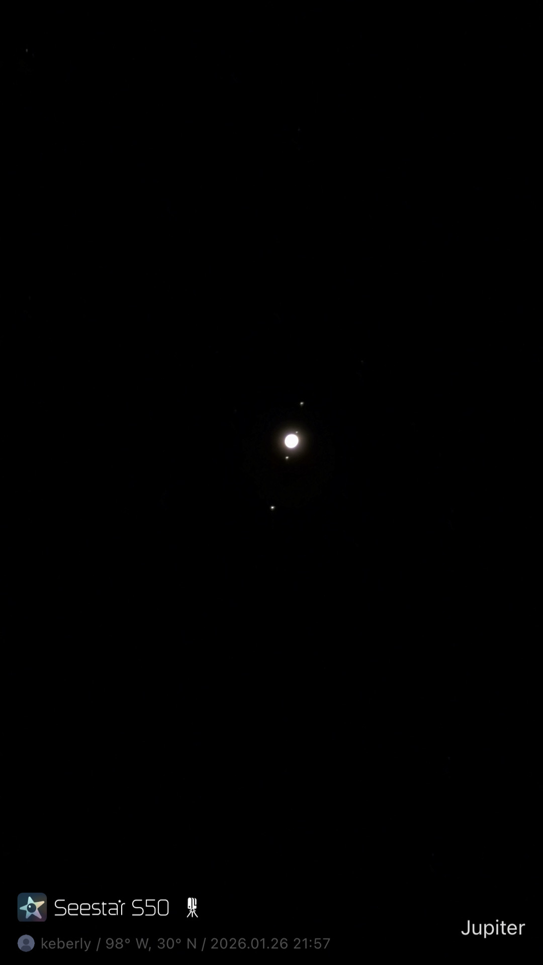 Jupiter and its four largest moons photographed from Lakeway, Texas with a ZWO Seestar S50 — Jupiter overexposed as a bright central point with three or four small bright dots (the Galilean moons) arranged around it against a black sky.