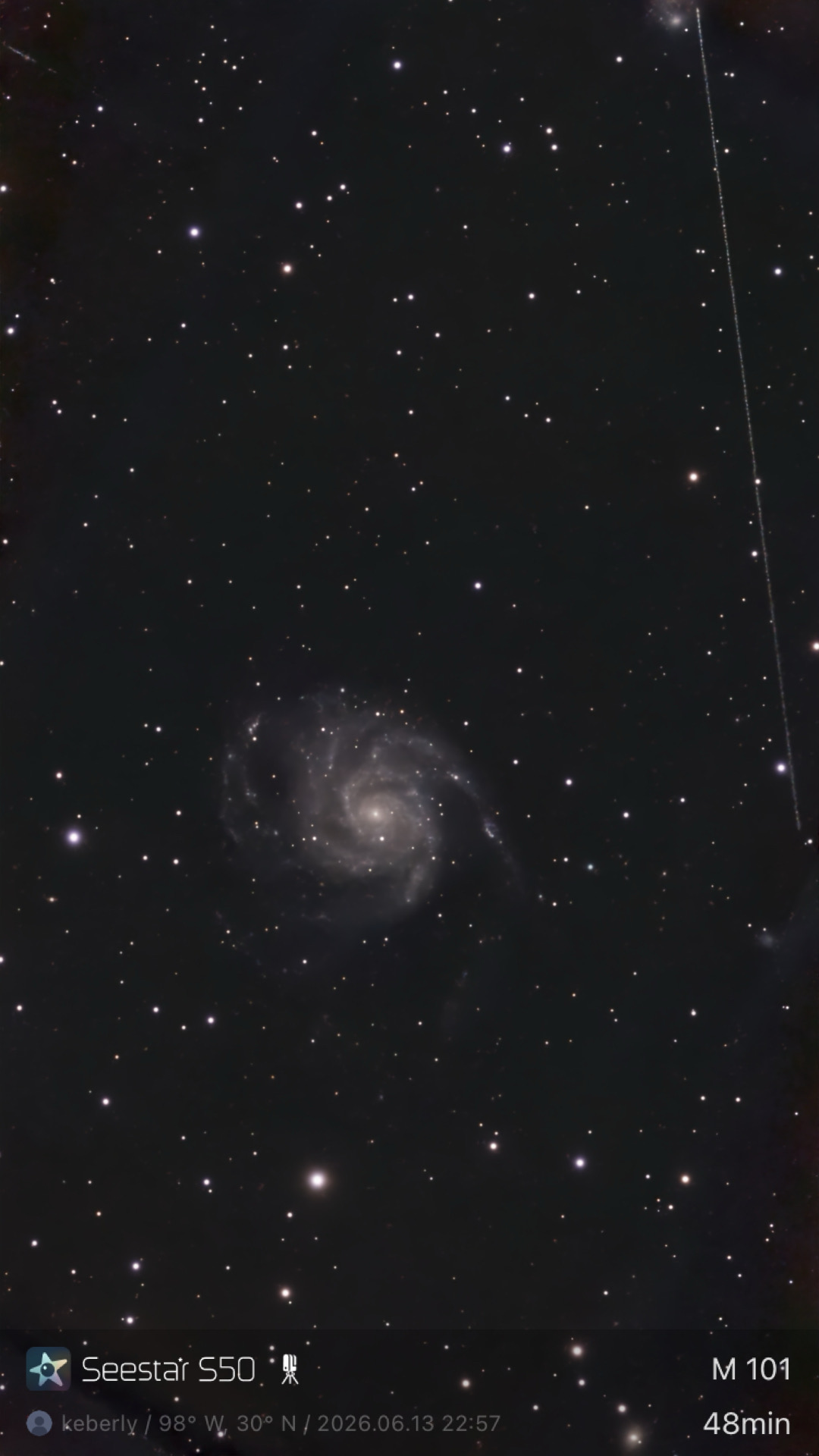 The Pinwheel Galaxy (M101) photographed from Lakeway, Texas with a ZWO Seestar S50 — a large face-on spiral galaxy with multiple resolved arms and star-forming regions, set against a dense starfield. A faint satellite trail cuts across the upper right of the frame.