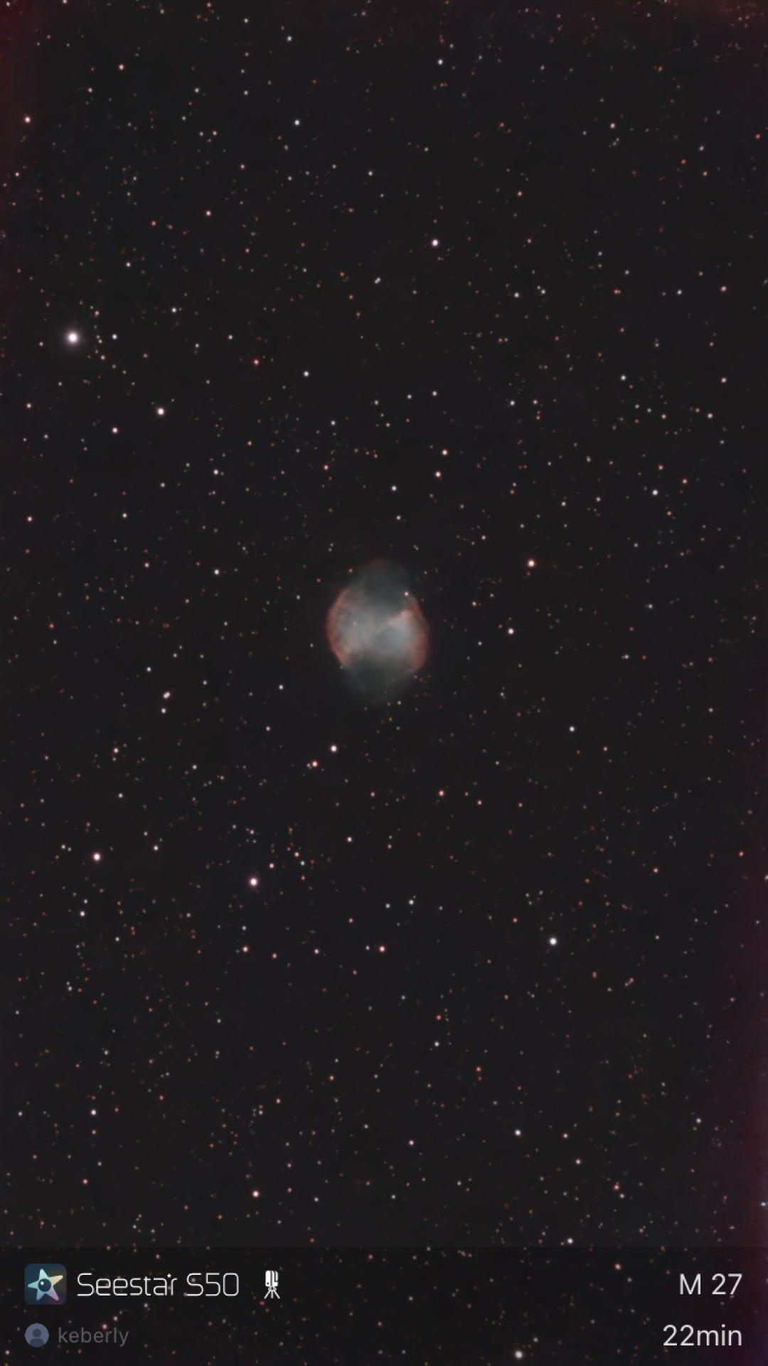 The Dumbbell Nebula (M27) photographed from Lakeway, Texas with a ZWO Seestar S50 — a small planetary nebula with a cyan core and pink/red outer lobes shaped like an apple core, against a dense starfield.