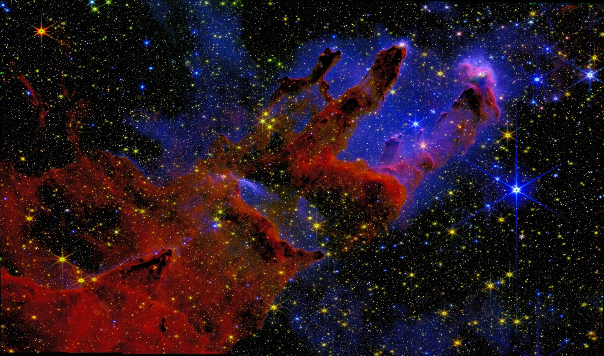 The Pillars of Creation in the Eagle Nebula (M16) — a JWST NIRCam image reprocessed by Kyle Eberly. Towering rust-red columns of gas and dust rise diagonally against a deep-blue, densely starred sky, with crimson glints of newborn stars at the pillar tips.
